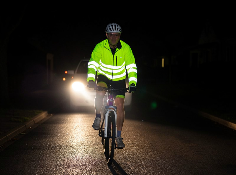 A cyclist in a fluorescent jacket with retroreflective panels lit up by car headlights on a dark Nordic street
